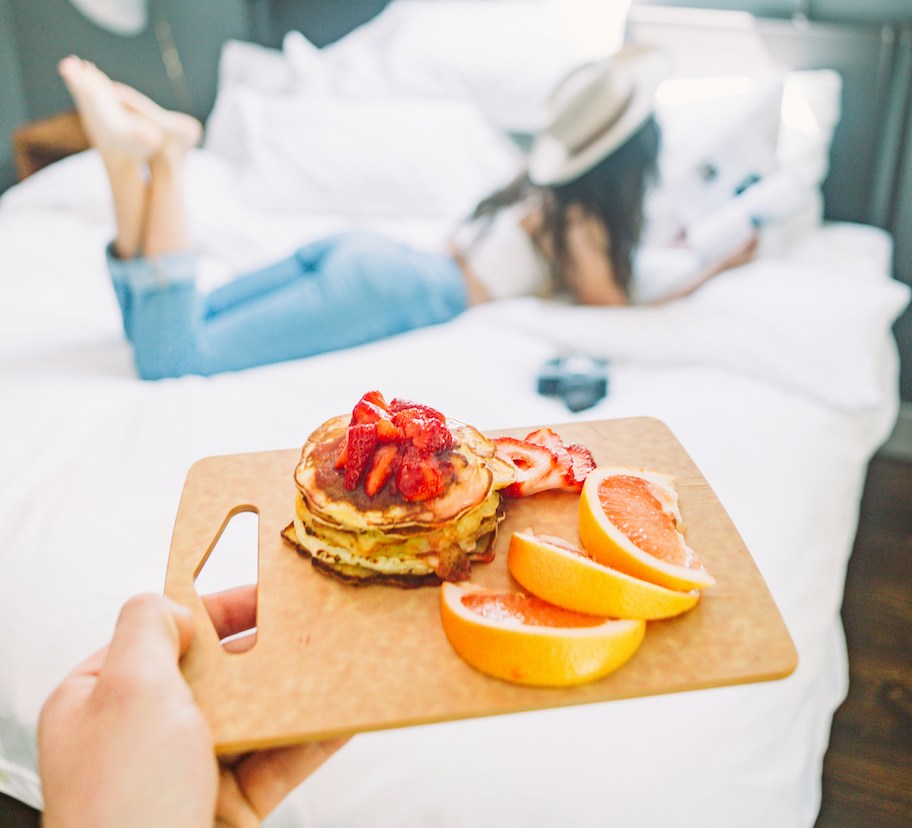 hand holding a cutting board with breakfast pancakes strawberries and woman laying on bed