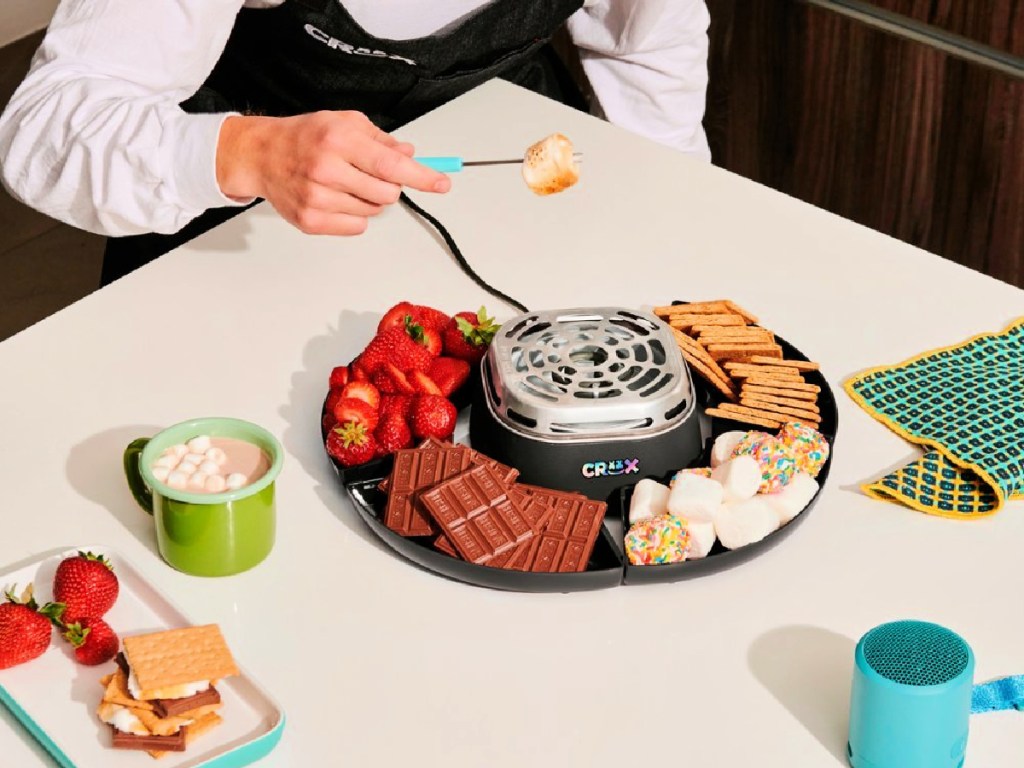 man in costume roasting a marshmallow over a crux smores maker filled with chocolate bars, marshmallows, strawberries and graham crackers