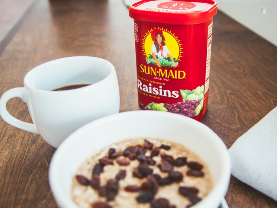sun maid raisins canister behind a bowls of oatmeal topped with raisins and a white mug full of black coffee