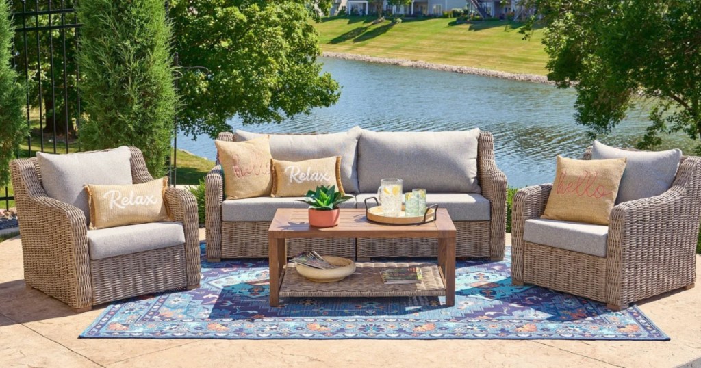 Wicker sofa, and two club chairs and coffee table outside on outdoor rug in front of river