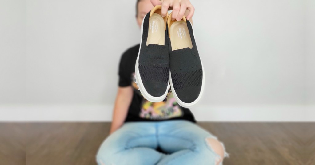 woman sitting on floor holding up pair of Dearfoams Sneakers in black