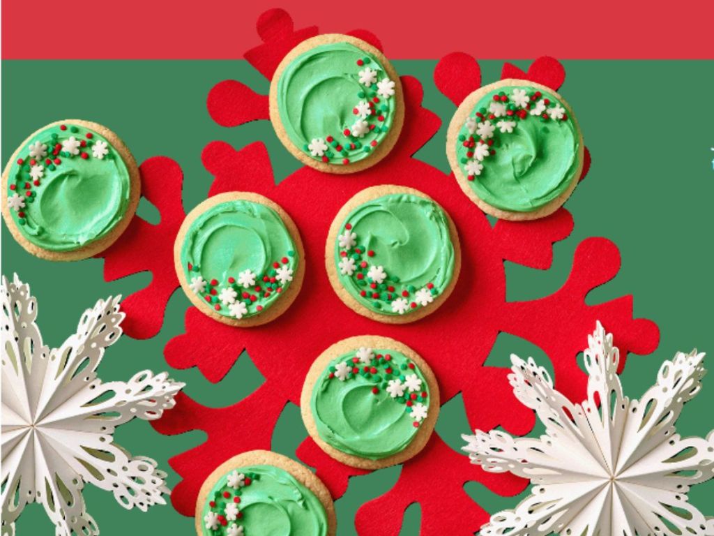 7 cookies with green icing and sprinkles on a red and green table cloth surrounded by paper snowflakes 
