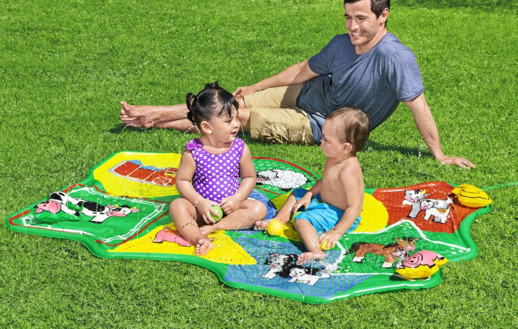 man sitting in grass with babies playing in barnyard themed splash pad
