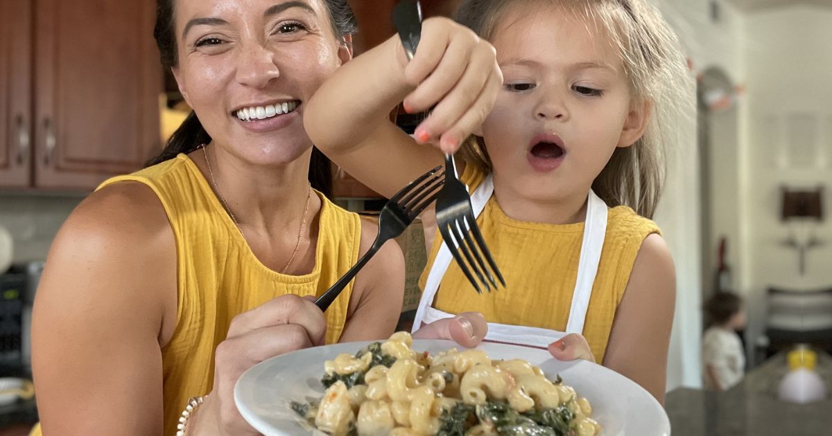 Little girl and mom holding a plate of pasta with little girl trying to dig into the pasta