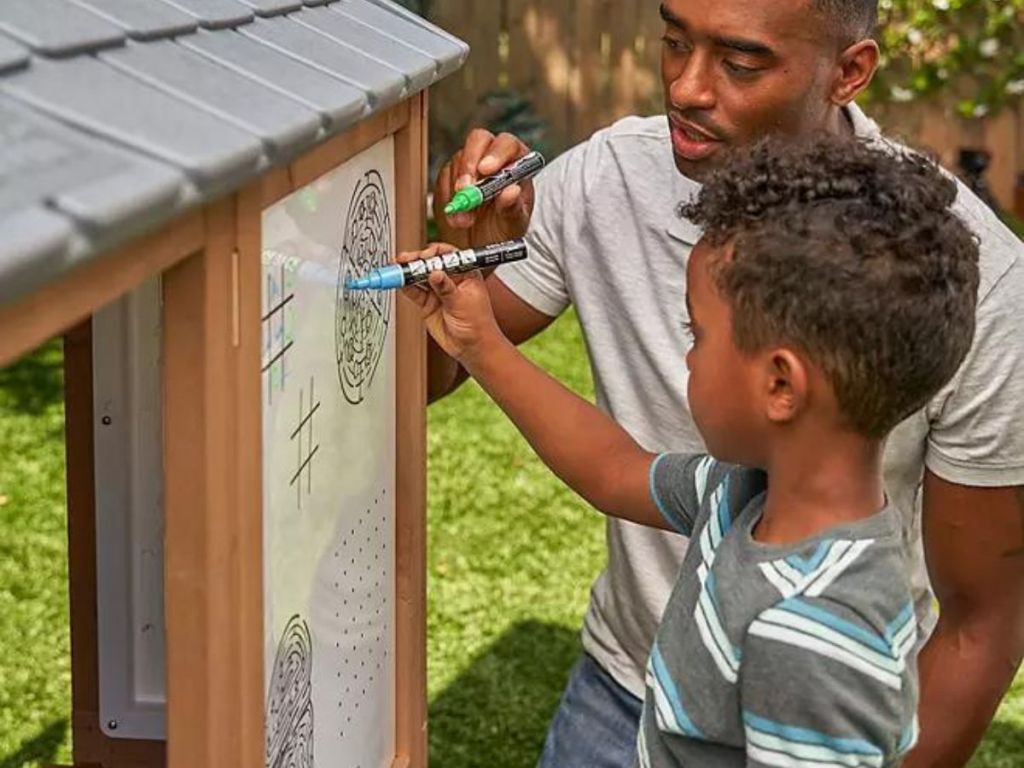Child writing on the whiteboard of a KidKraft Grill & Chill Pizza Party Wooden Outdoor Playhouse