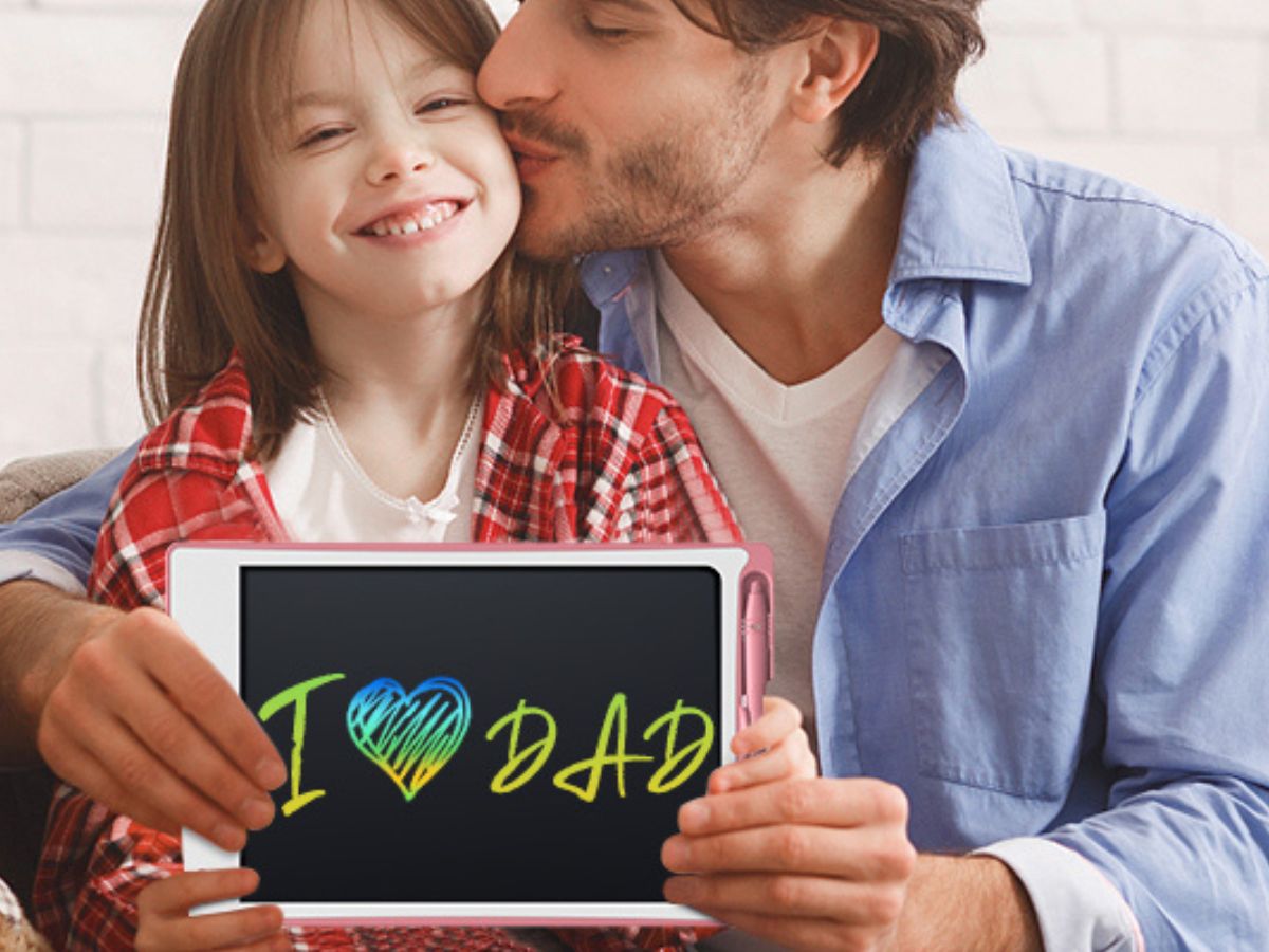 A child holding an erasable board that says "I love Dad" while their father is kissing their cheek. 