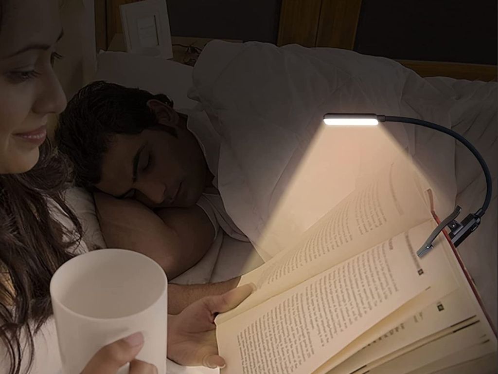 A woman reading in bed with a book light while a man sleeps 