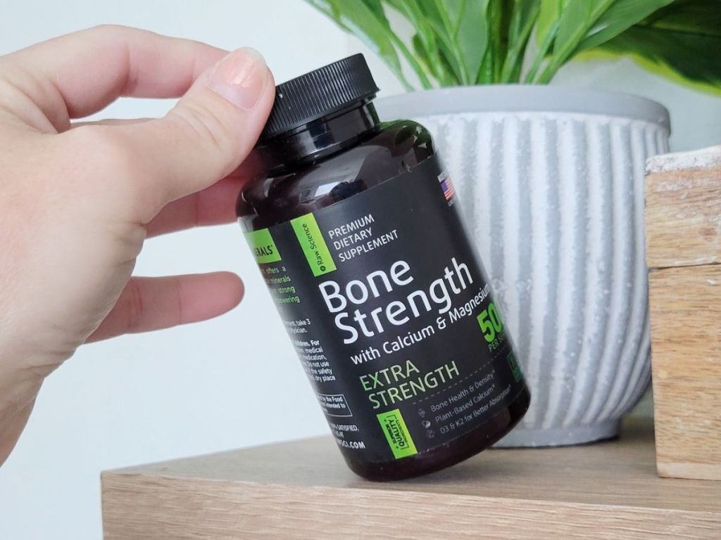 Raw Science Bone Strength Supplement on a shelf next to a plant with the supplement bottle being slightly tipped by a hand