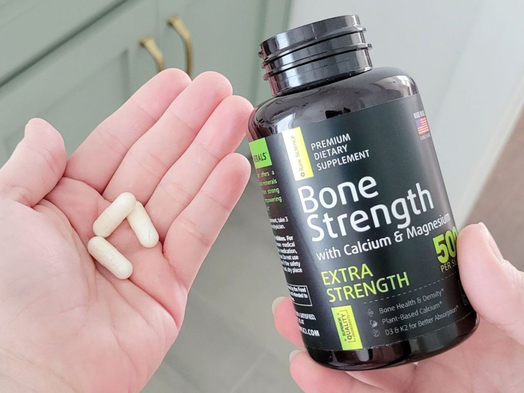 One hand holding a bottle of Raw Science Bone Strength supplements and an open hand next to it with 3 capsules in the palm.