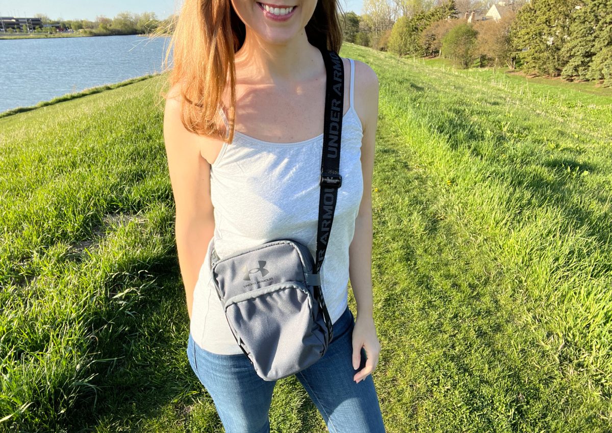 smiling woman wearing an under armour crossbody bag, jeans, and a tank top, standing in grass near water