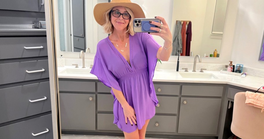 woman taking mirror selfie wearing purple swim cover up with beach hat