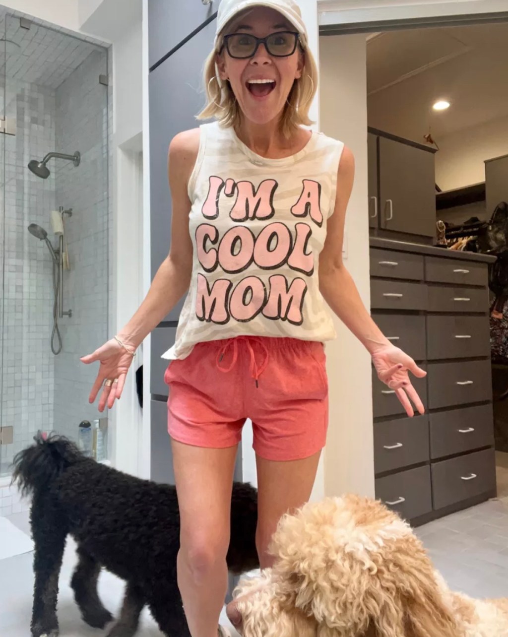 woman wearing i'm a cool mom shirt with pink shorts and hat standing in bathroom with two dogs