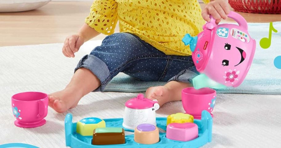 child in yellow shirt playing with pink teapot set on floor