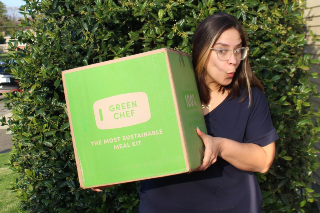 woman with surprised face holding green chef box next to bush