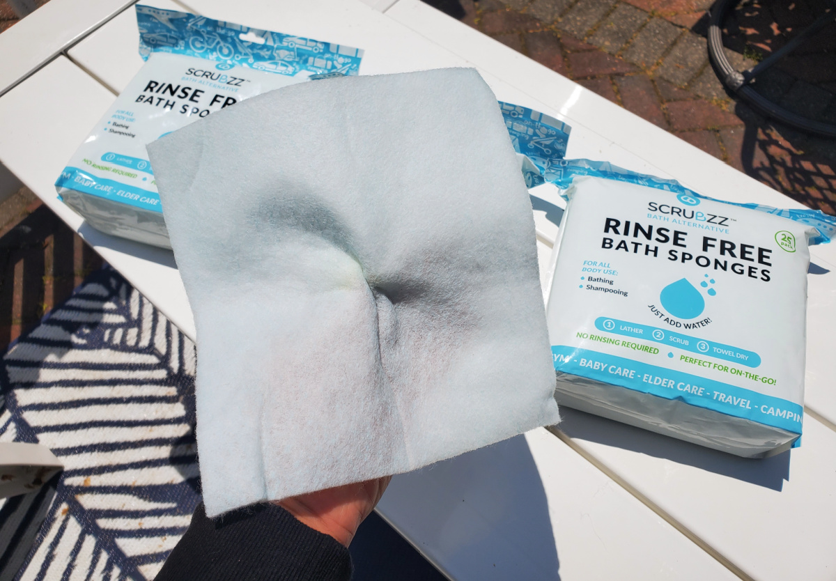 hand holding a scrubzz rinse free bath sponge sheet above other packages of the product lying on a table