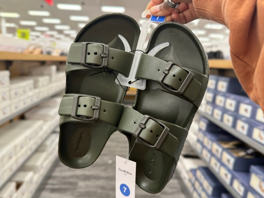 hand holding up a pair of men's footbed sandals in olive green in a store aisle