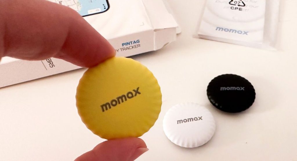 MOMAX Tracker Tag shown in all 3 colors, woman holding the yellow one, box in the background