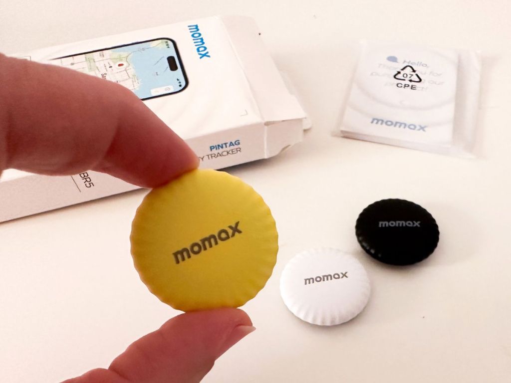 MOMAX Tracker Tag shown in all 3 colors, woman holding the yellow one, box in the background