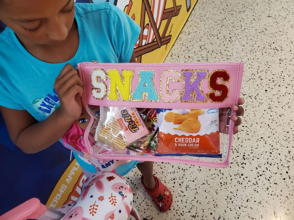 Little girl holding a Chenille Letter Clear Makeup Bag Purse - Stoney Clover Lookalike that says "Snacks" and has snacks in it
