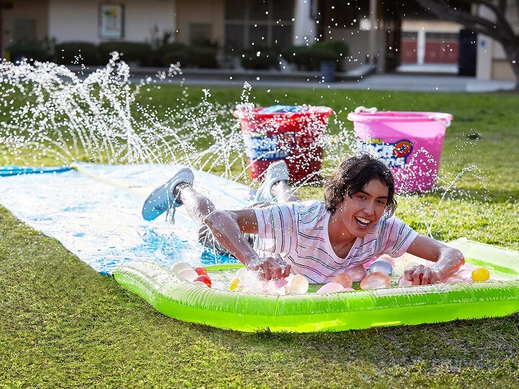 guy going down a lawn water slide with water balloons at the end