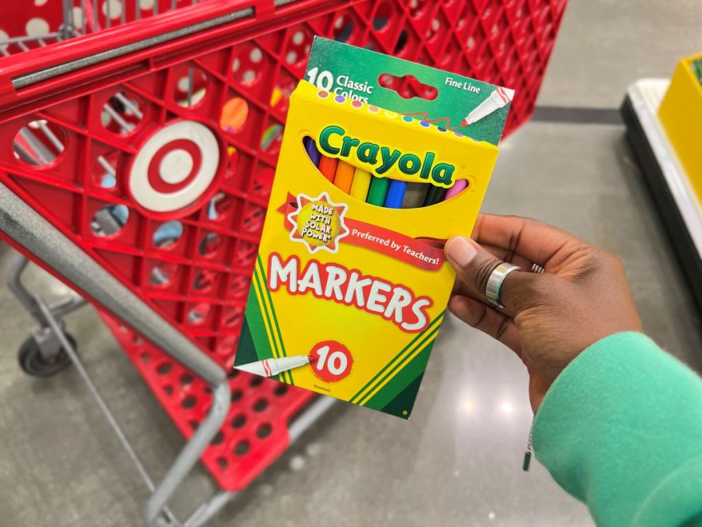 Hand holding a pack of Crayola markers next to a Target cart