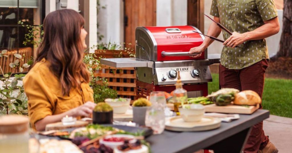 a woman at an outdoor table with food on it and a man using a 2 burner grill in the background 