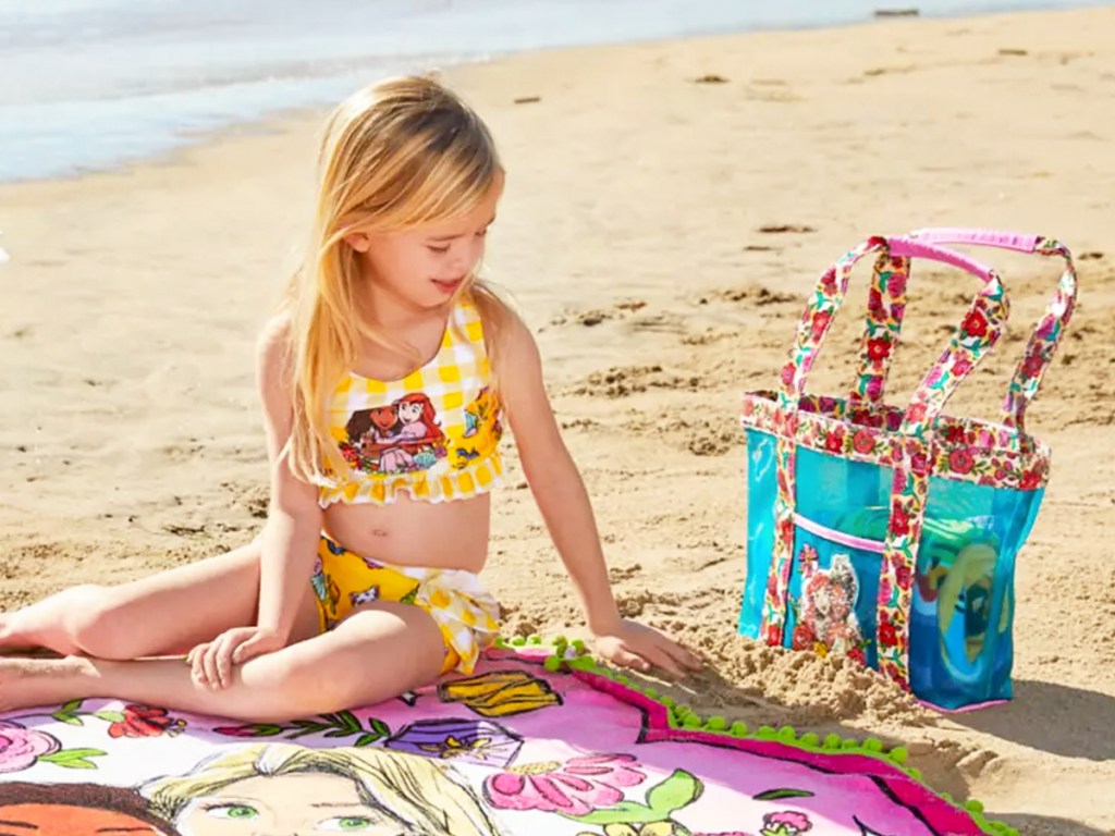 girl sitting on towel at beach next to a beach bag