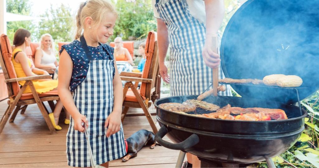 Family grilling on a charcoal grill with a dad and little girl in blue and white aprons