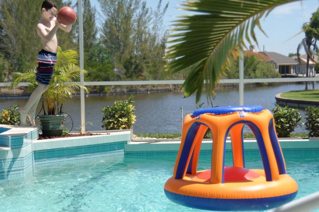 Teenage boy with a basketball jumping into a pool and shooting the ball into a SWIMLINE Inflatable Pool Basketball Hoop Floating Or Poolside Game 