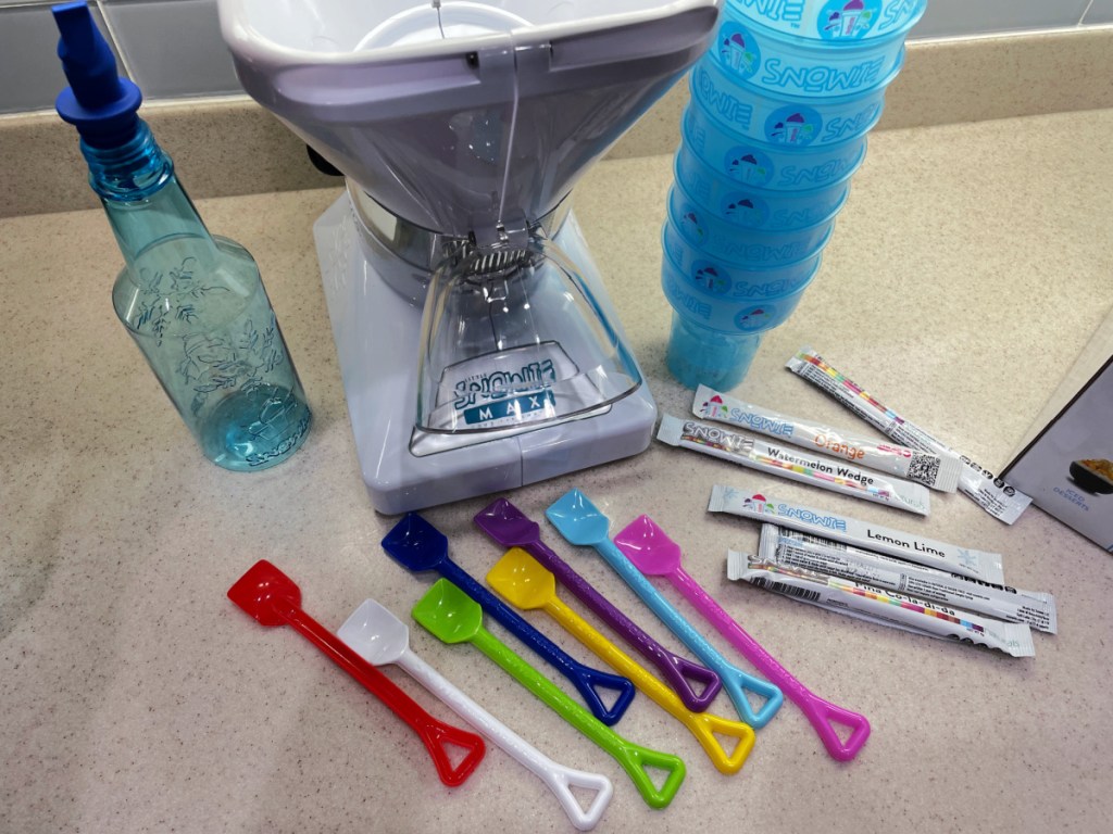 Little Snowie MAX Shaved Ice Machine sitting with syrup bottles, cups, spoons, and flavorings on a countertop