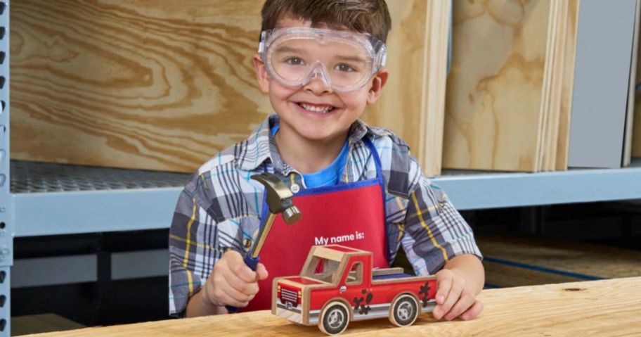Little boy wearing safety goggles and holding a hammer in one hand and a wooden pickup truck in the other