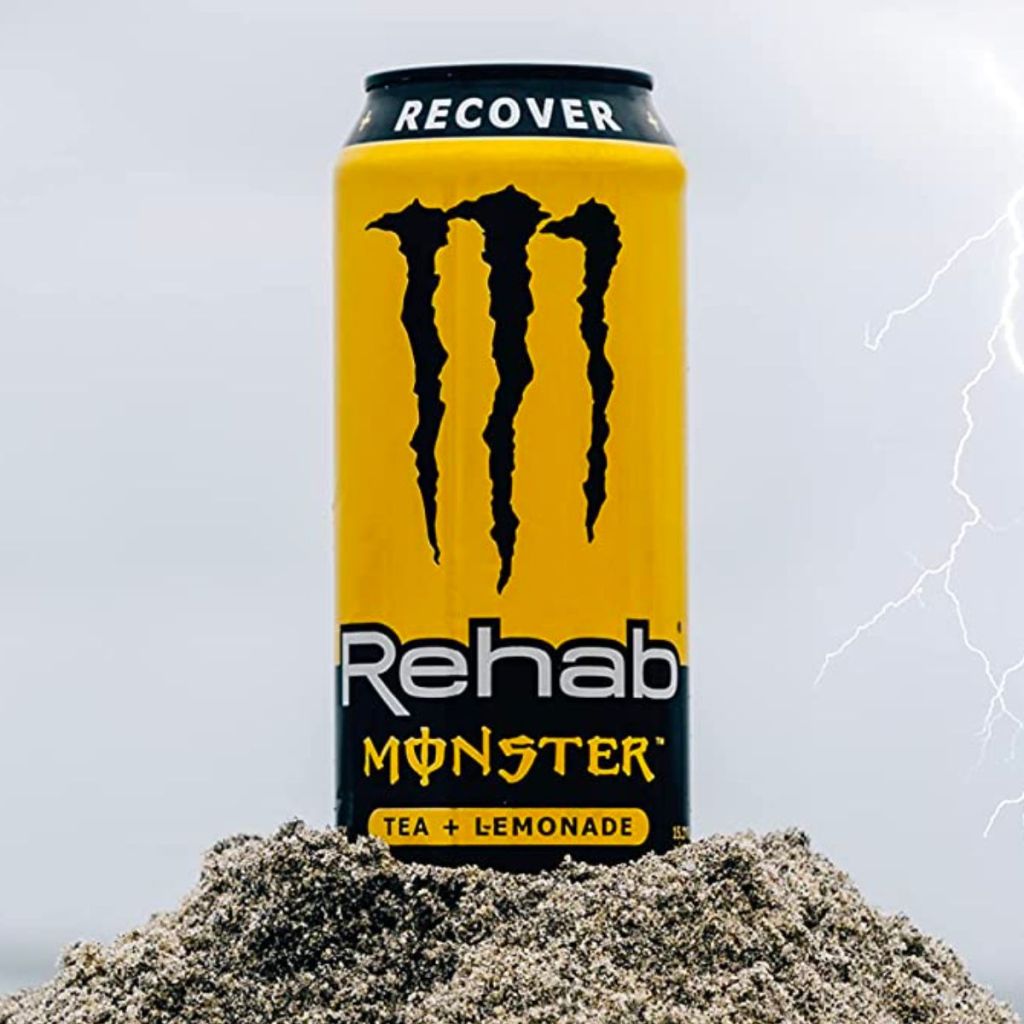 a can Monster rehab tea + lemonade in close up sitting on a pile of wet beach sand