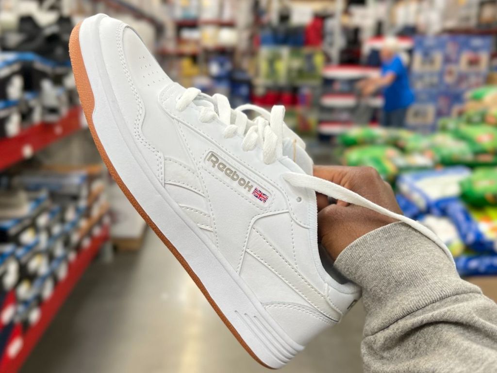 A hand holding a white Reebok sneaker in a store