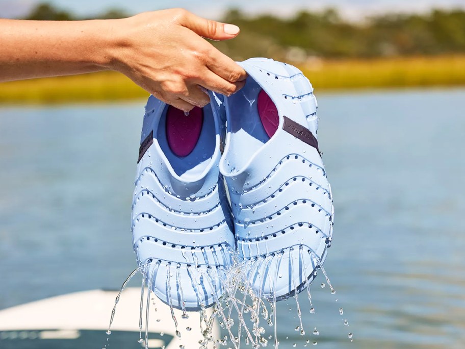 person holding up a pair of light blue water shoes with water draining out of them