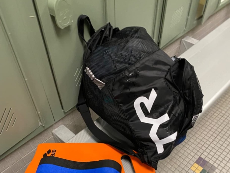 40L TYR mesh mummy backpack in black leaning against lockers with orange kick board