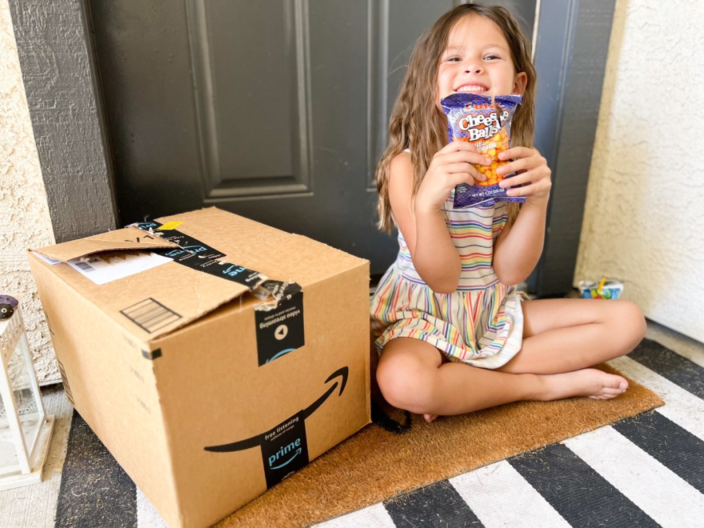 girl at front door sitting next to amazon box with snack in hands