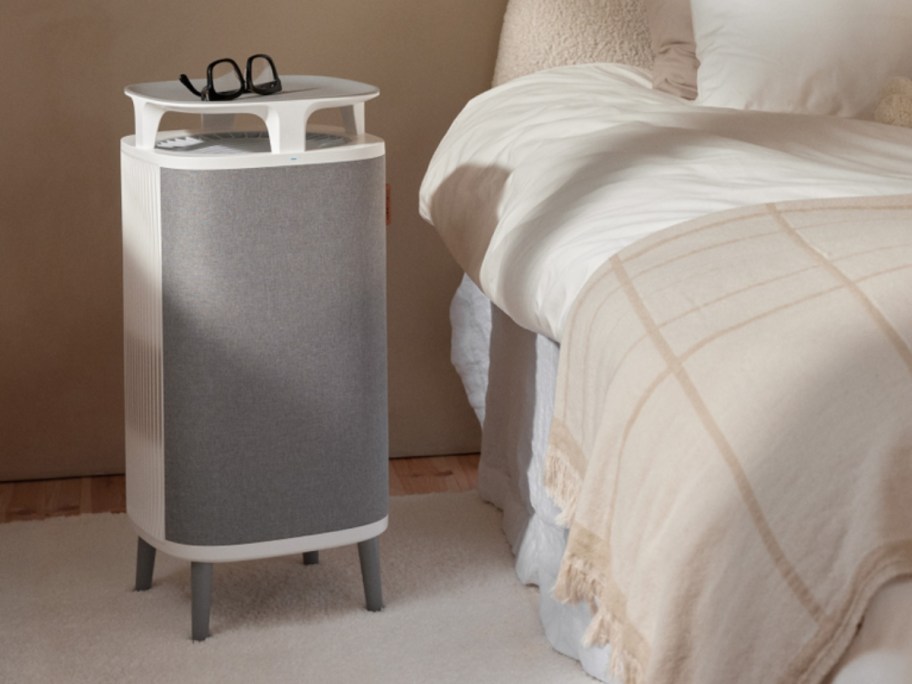 a large grey and white air purifier machine with feet next to a bed, a pair of eye glasses sits on of the air purirfier