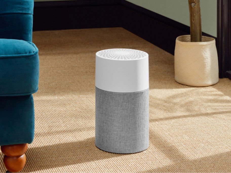 a small/ medium sized grey and white air purifier sitting in a living room floor by a blue chair, potted plant behind it