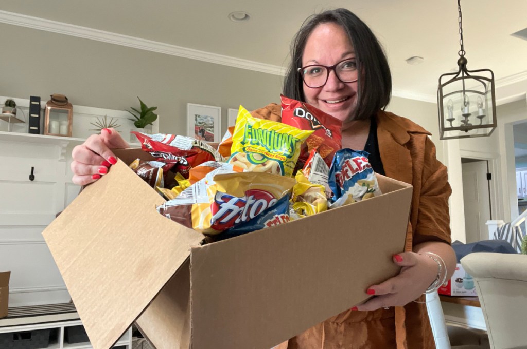 smiling woman holding a huge amazon box filled with frito lay chips in her living room