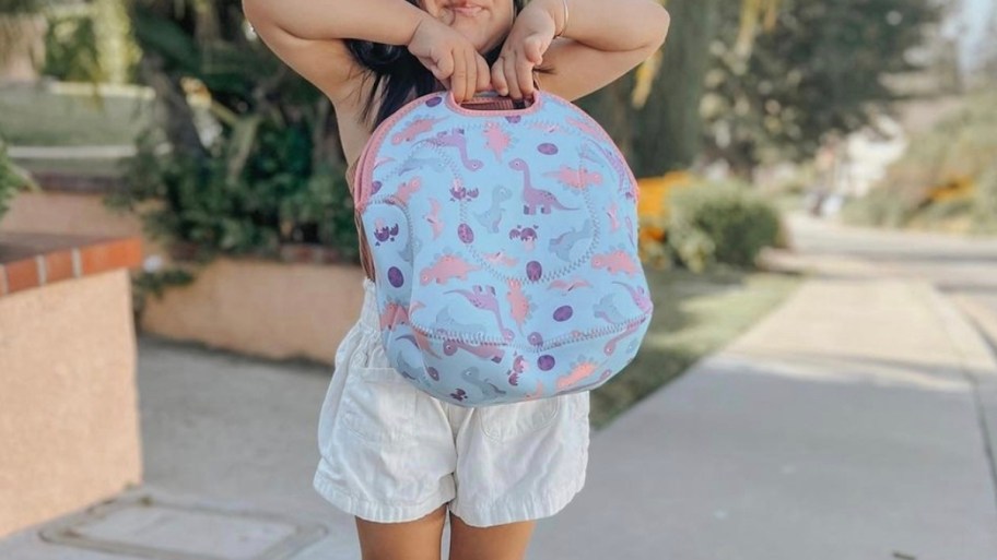 girl holding up dinosaur lunch bag outside on sidewalk