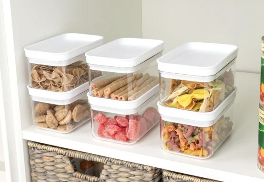 six small clear and white food storage containers with snacks inside on a pantry shelf