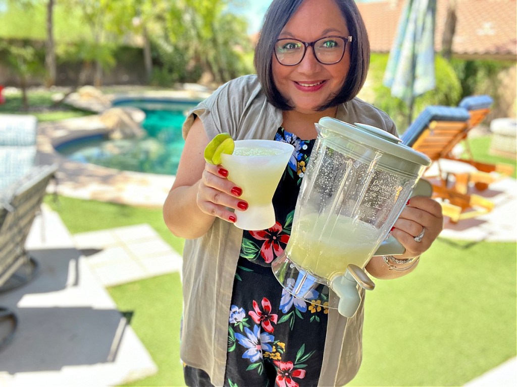 woman standing in a sunny backyard, holding a slushie maker and a plastic glass, both filled with frozen margaritas