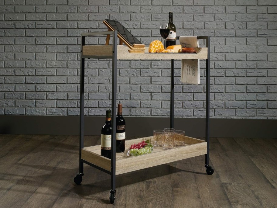 wood and metal bar cart with food and wine bottles on it sitting in a room with a wood floor and white brick wall
