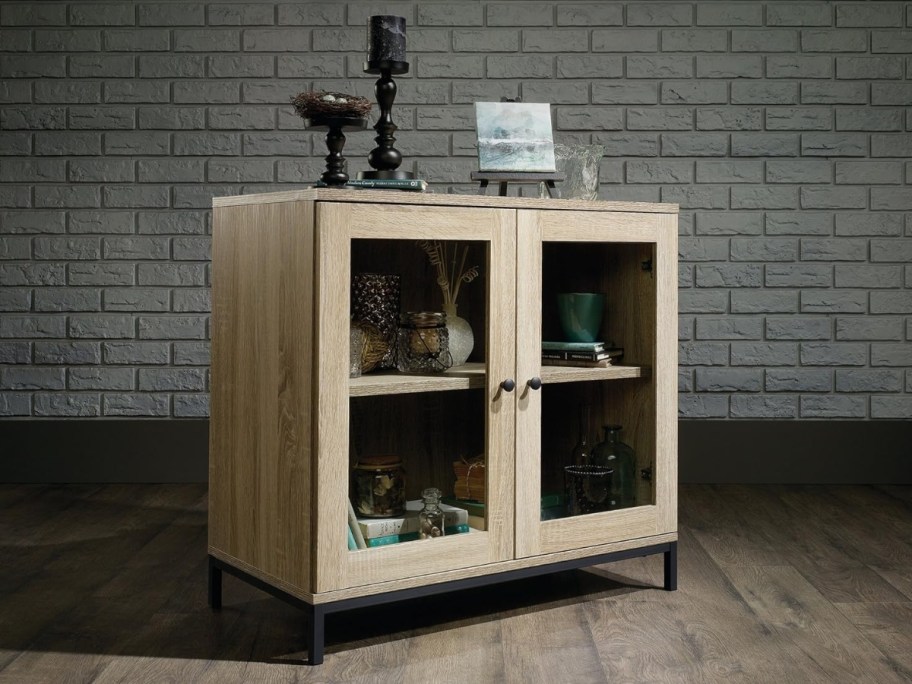 oak color display furniture case with glass doors, metal stand sitting in a room with a wood floor and white brick wall