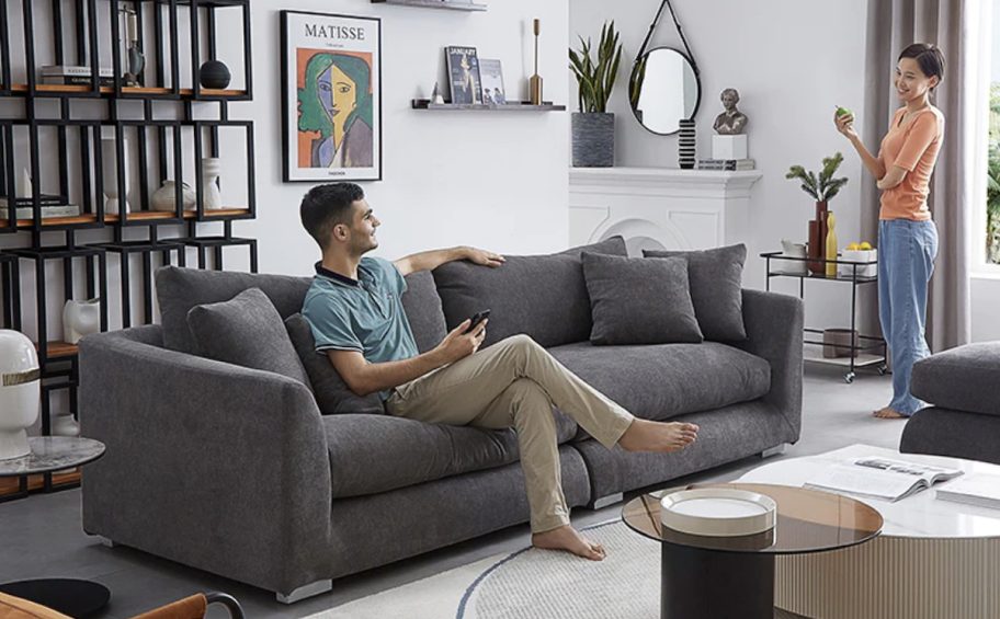 man sitting on gray couch in styled living room talking to woman holding pear