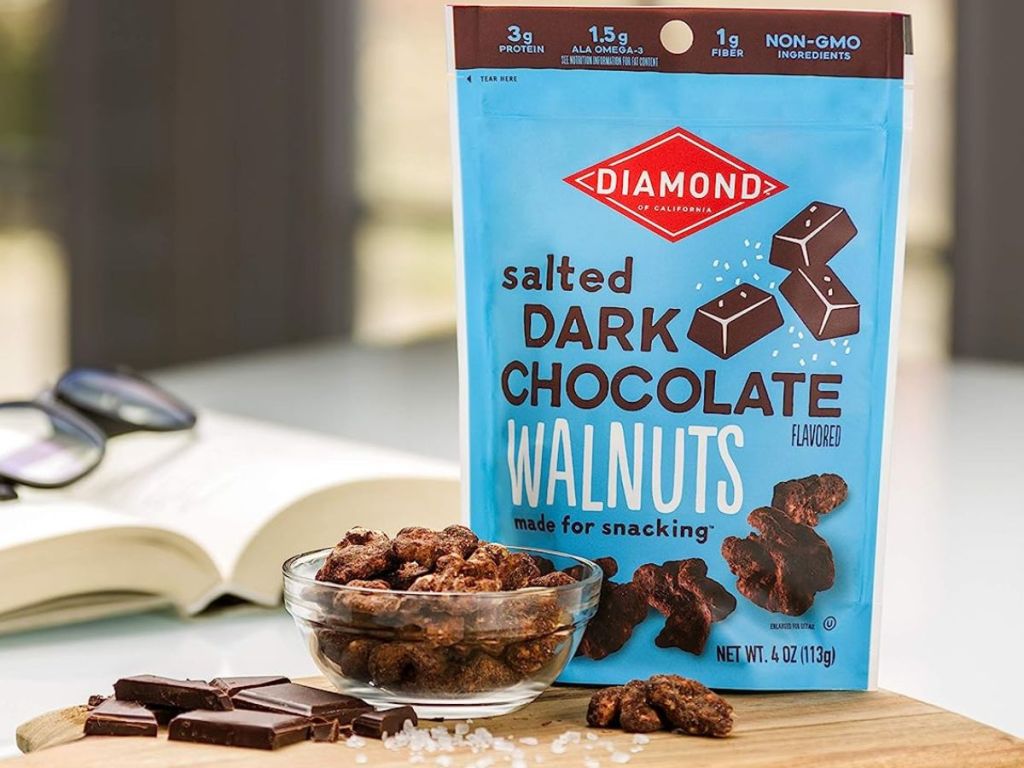 Diamond of California Salted Dark Chocolate Walnuts next to a bowl of nuts and a book with glasses on it