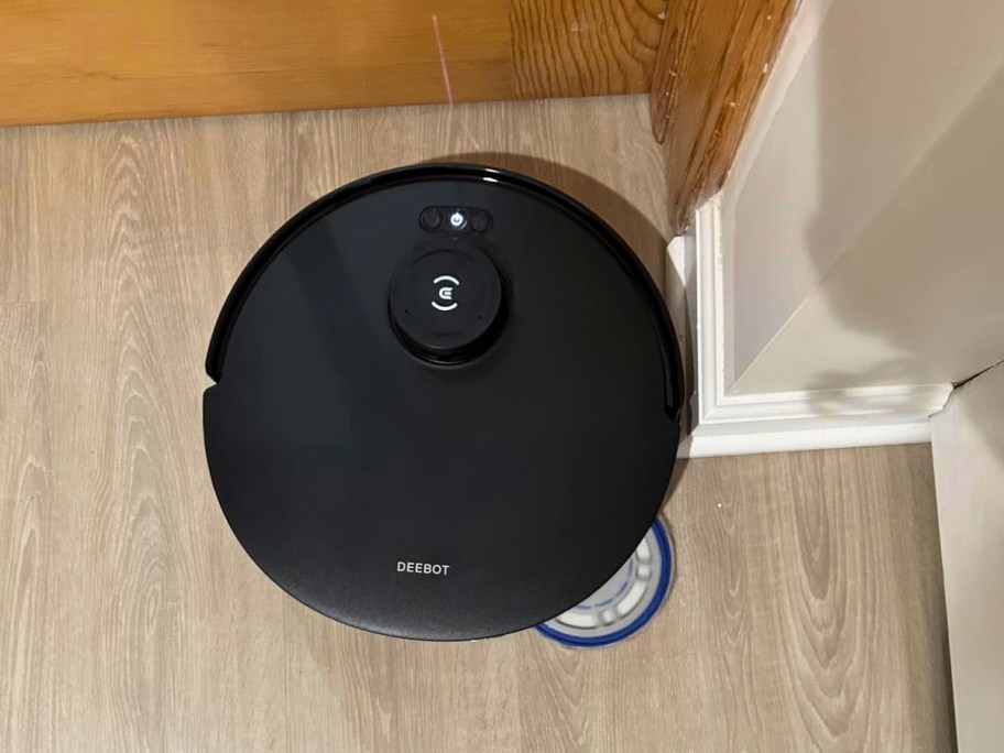 black Ecovacs Deebot Robot Vacuum & Mop cleaning into the corner of a room on a hardwood floor.