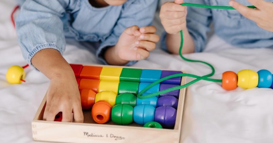 two kids laying on a bed playing with a set of wooden primary color beads and green lacing strings in a wooden box that says 