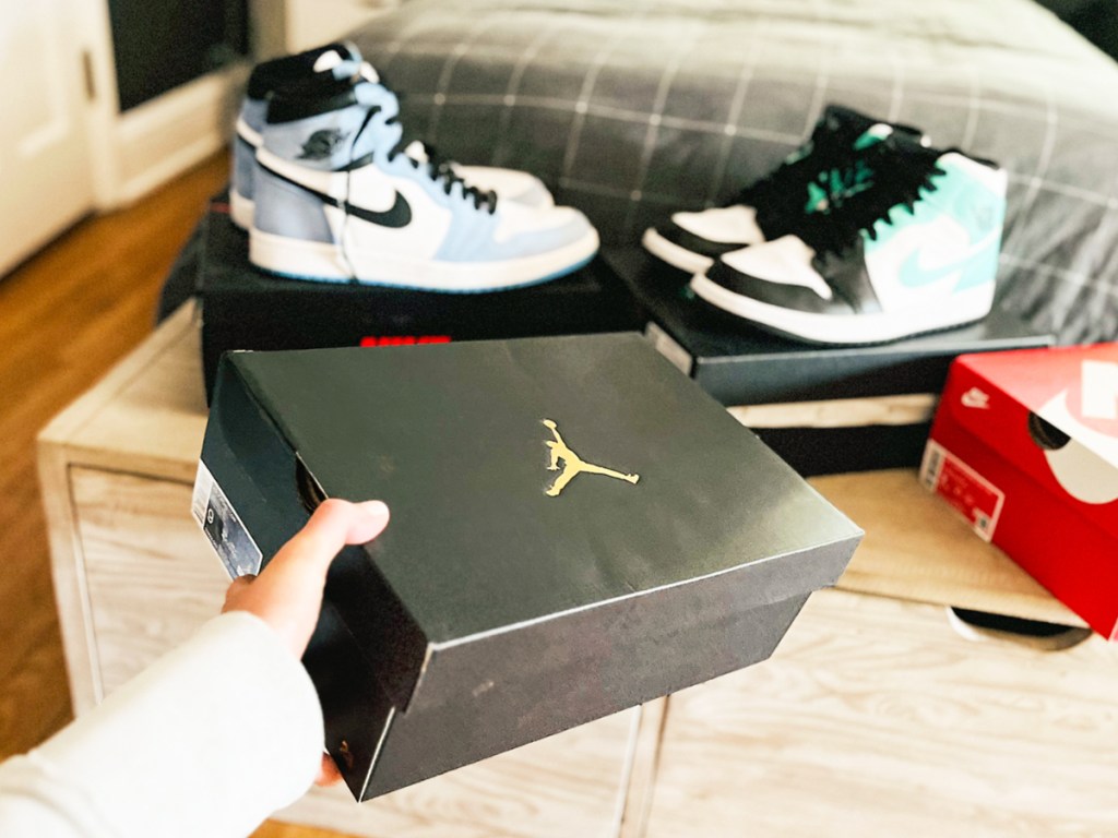 hand holding a black jorden shoe box with two pairs of shoes in the backpground