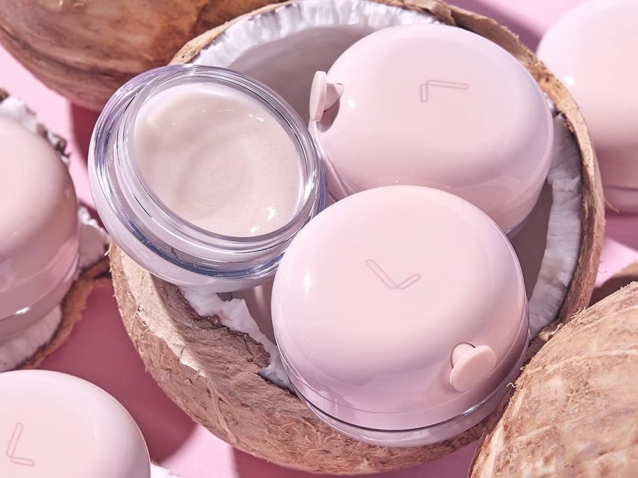 3 jars of light pink lip cream in a coconut shell, one open showing cream, with others around it